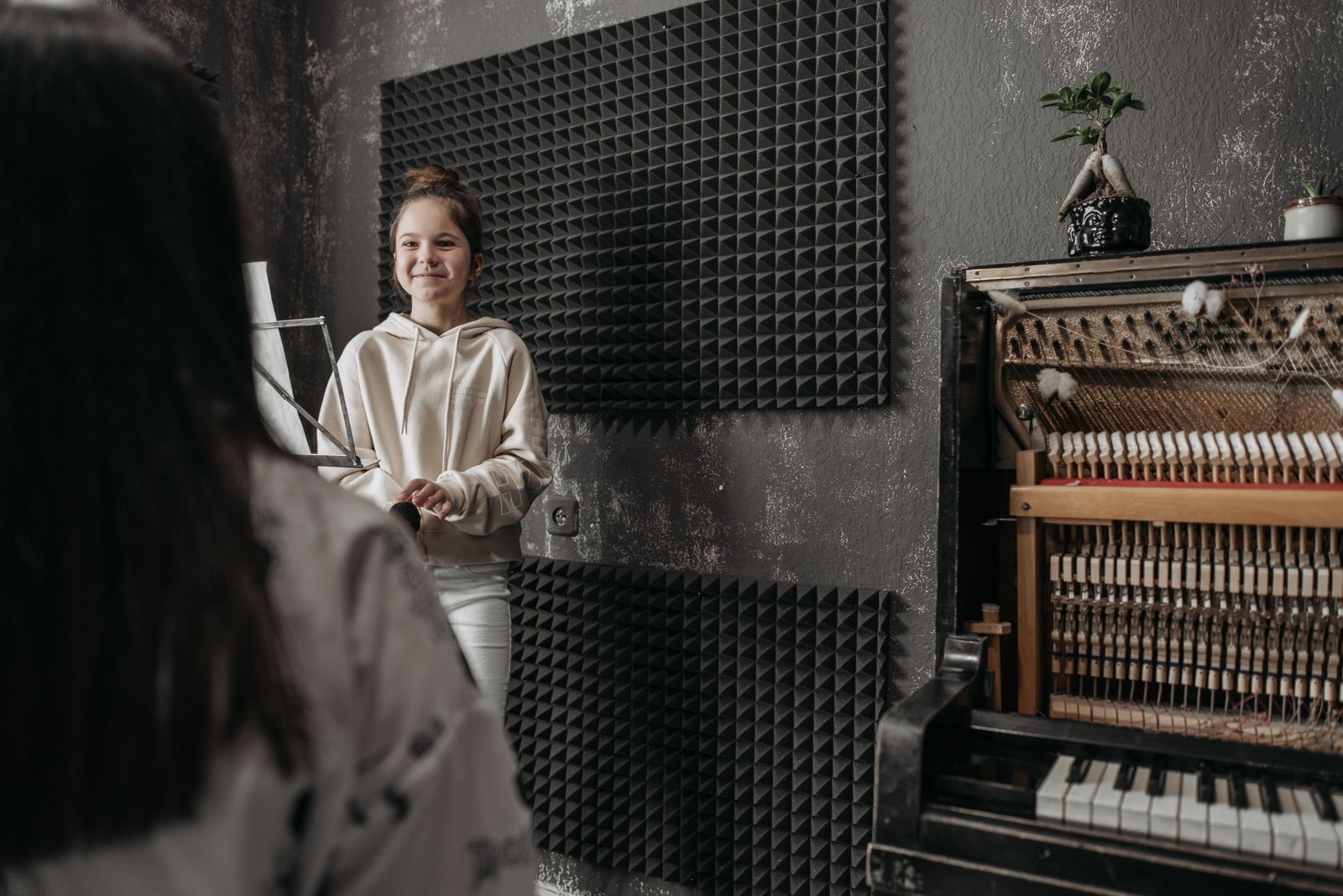 Aluna sorrindo dentro do estúdio da escola, diante de um piano vertical aberto, durante a aula de canto.