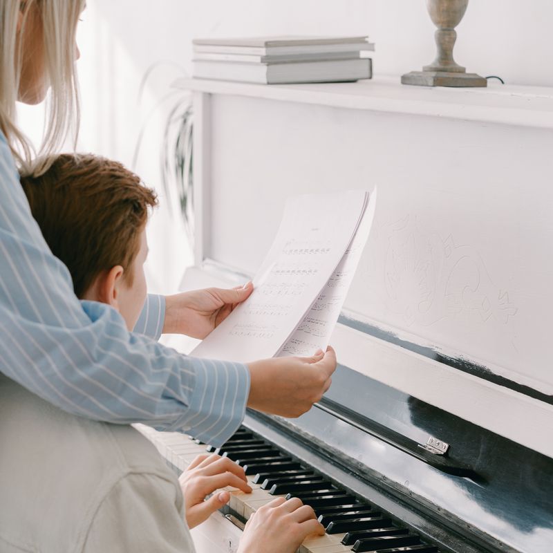 Adulto e criança sentados juntos ao piano, lendo a mesma partitura enquanto a criança toca.