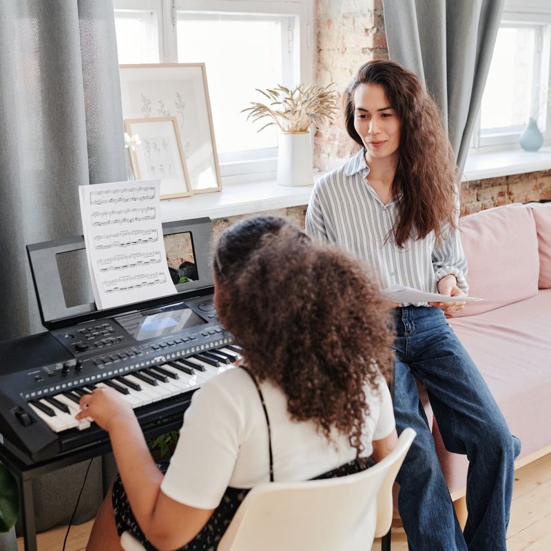 Mulher sentada com a partitura na mão acompanha uma aluna ao teclado numa sala de aula da escola.